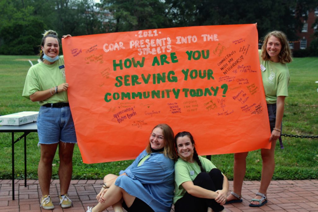 COAR staff posing with a paper banner that says "2021 COAR Presents Into the Streets: How are you serving your community?" There are many handwritten responses on the banner, including "connecting with my neighbors" and "appreciating Mother Earth."