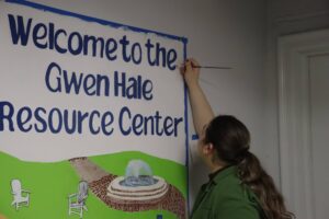 Person painting part of a mural that says "Welcome to the Gwen Hale Resource Center." 