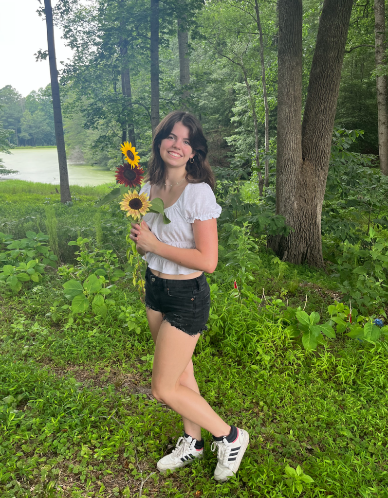 Hope McCallister with brown hair, a white shirt, holding sunflowers outside.