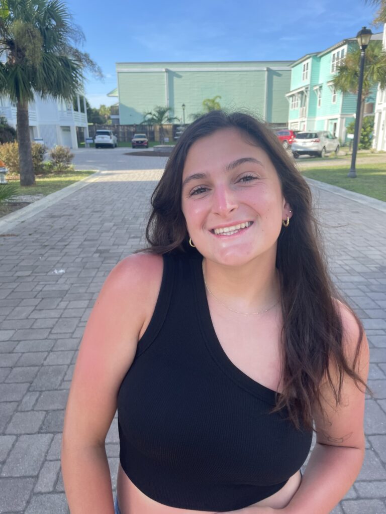 Victoria, with dark hair, wearing a black tank-top, standing in front of palm trees.