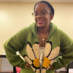 Martina, wearing glasses and a green sweater with a daisy on it.