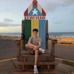 Everett wearing a gray shirt, black shorts, and white shoes while sitting on a colorful chair near the ocean.
