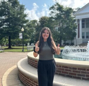 Picture of a young woman with brown hair standing in front of a fountain giving the "thumbs up" symbol. 