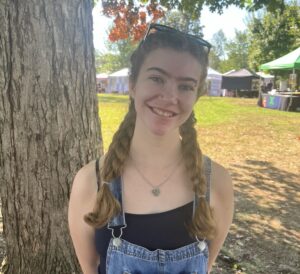 Jaydn stands in front of a tree wearing a black sleeveless top with blue denim overalls. Her hair is in two braids on either shoulder and she is wearing a necklace.