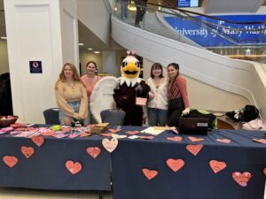 Four people standing in a line with Sammy D. Eagle mascot in the middle. Sammy has wings and a heart headband on. There are hearts plastered all over the front of the tables they are standing behind, which are covered in a navy blue table cloth.