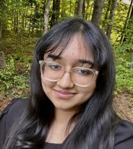 Aliya smiling in a black dress shirt, standing in the woods.
