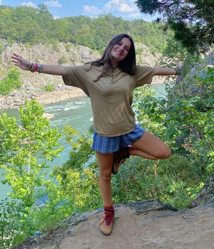 Eva standing on one leg with both arms out in front of a scenic river background. She is wearing a tan tee shirt, patterned blue shorts, hiking boots, and numerous bracelets.