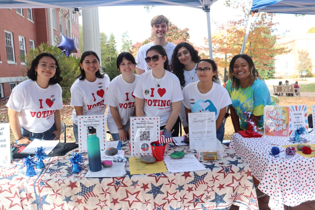Seven students stand behind a table wearing t-shirts that say "I love voting". 