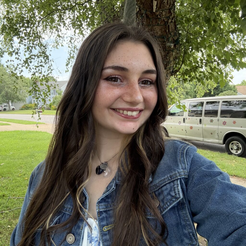 Sarah, wearing a denim jacket, smiling in front of a tree.